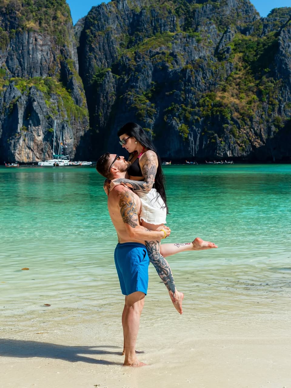 Tattooed man and woman embracing in turquoise tropical water with limestone cliffs in background, displaying visible arm and shoulder tattoos in black ink.