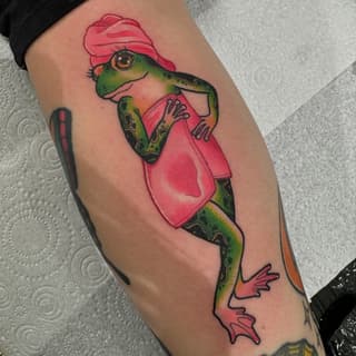 Colorful frog wearing a pink cap and jacket tattooed on the upper leg, featuring green, orange, and pink hues.