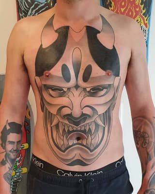 Large demon or oni mask face tattoo covering the chest in black and gray ink with shaded horns extending across the shoulders.