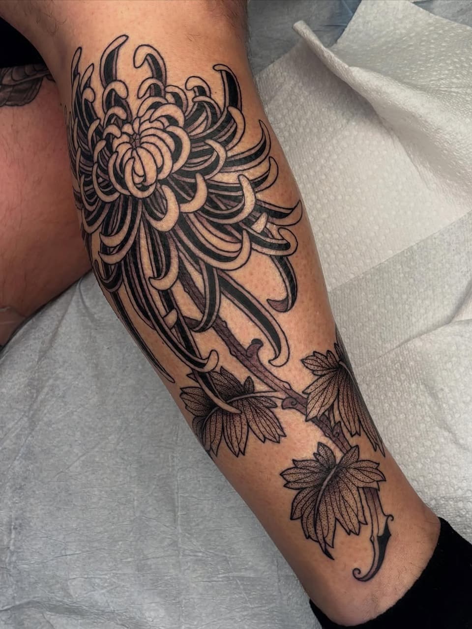 Detailed chrysanthemum flower tattoo on forearm with curved petals and decorative leaf elements below in black ink.