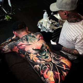 Tattoo artist applying colorful floral and decorative back piece featuring red, orange, and green flowers with ornamental scrollwork on client's back.