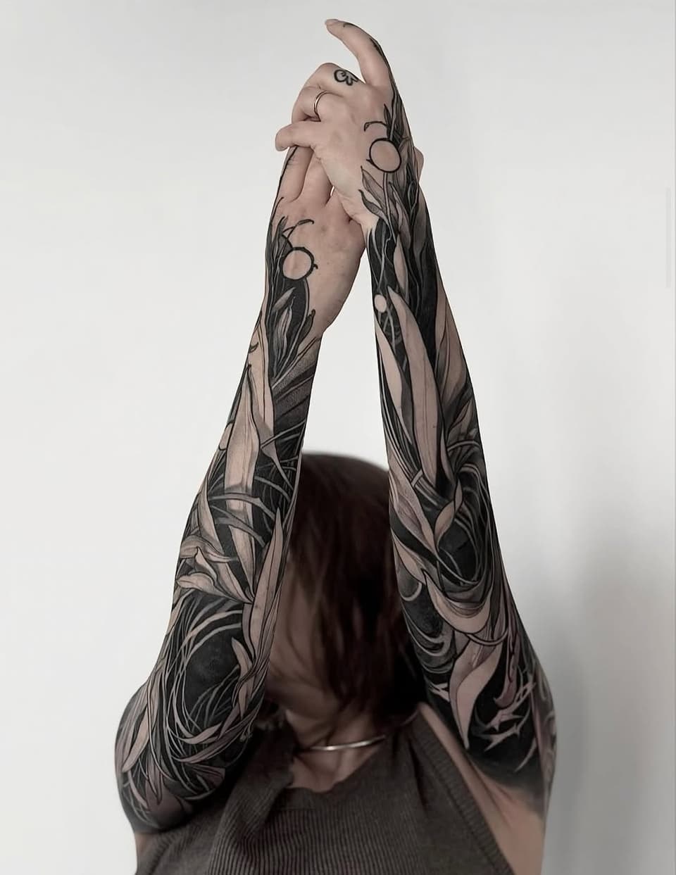 Person displaying full sleeve tattoos featuring botanical and circular motifs in black and gray ink on both arms, shown from behind.