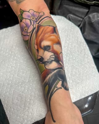 Colorful forearm tattoo featuring a fox head with flowers, decorative linework, and a bat or wing motif below in orange, purple, green, and black.