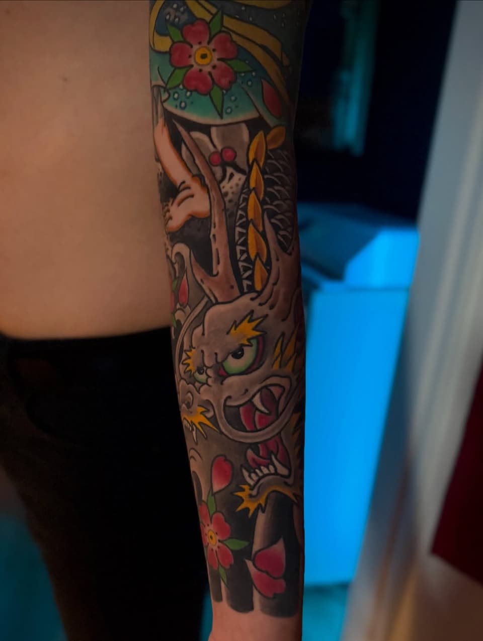 Colorful full-sleeve tattoo featuring floral motifs, dragon heads with spikes, and decorative elements in red, green, yellow, and brown on the forearm and arm.