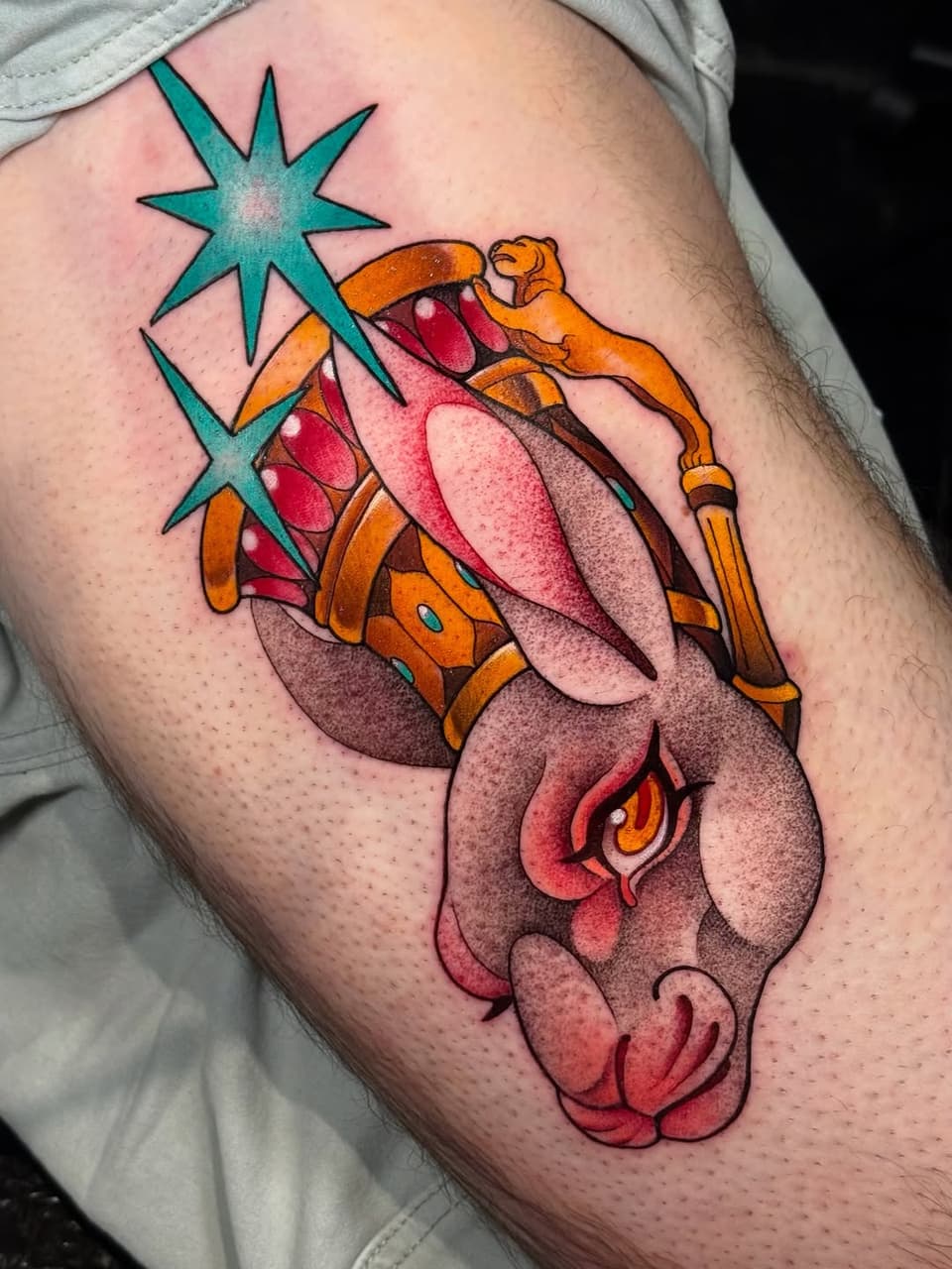 Colorful tattoo on thigh featuring a lion's head in orange and pink with a turquoise star above and a small monkey figure, highlighted with shading and details.