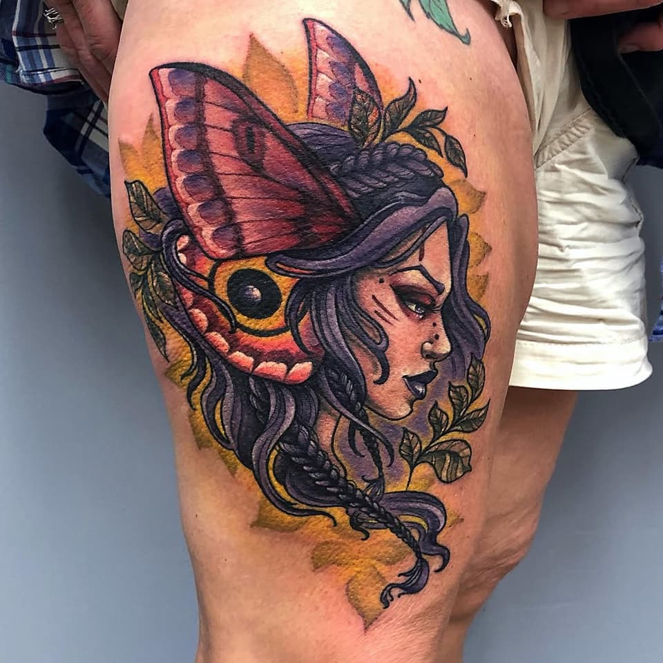 Colorful thigh tattoo featuring a woman's profile with a large butterfly wing headdress, surrounded by leaves and warm orange and purple tones.