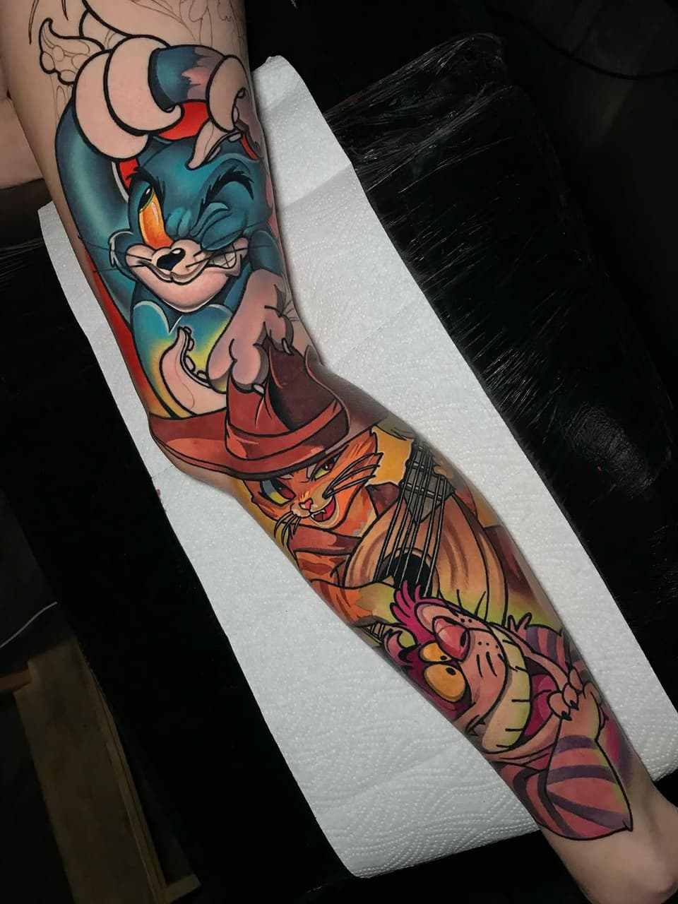 Colorful cartoon character tattoo on forearm featuring a blue cat and orange cat with red clothing, vibrant gradient backgrounds, and bold black outlines.