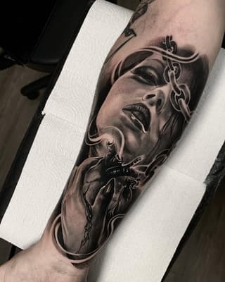 Detailed monochrome arm tattoo featuring a woman's face with ornamental chains and a heart with thorns, showcasing shading and portraiture work.