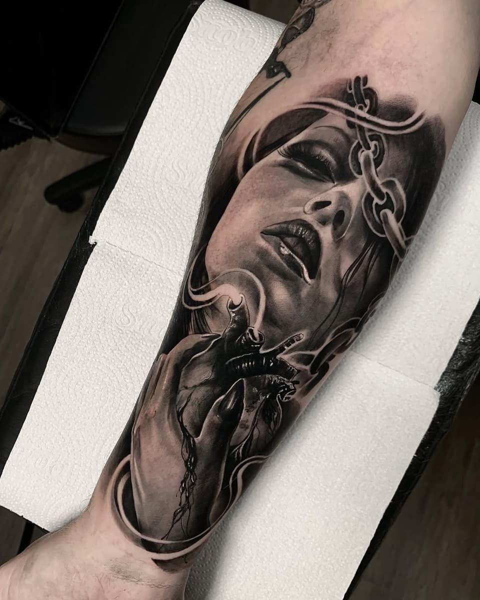 Detailed monochrome arm tattoo featuring a woman's face with ornamental chains and a heart with thorns, showcasing shading and portraiture work.