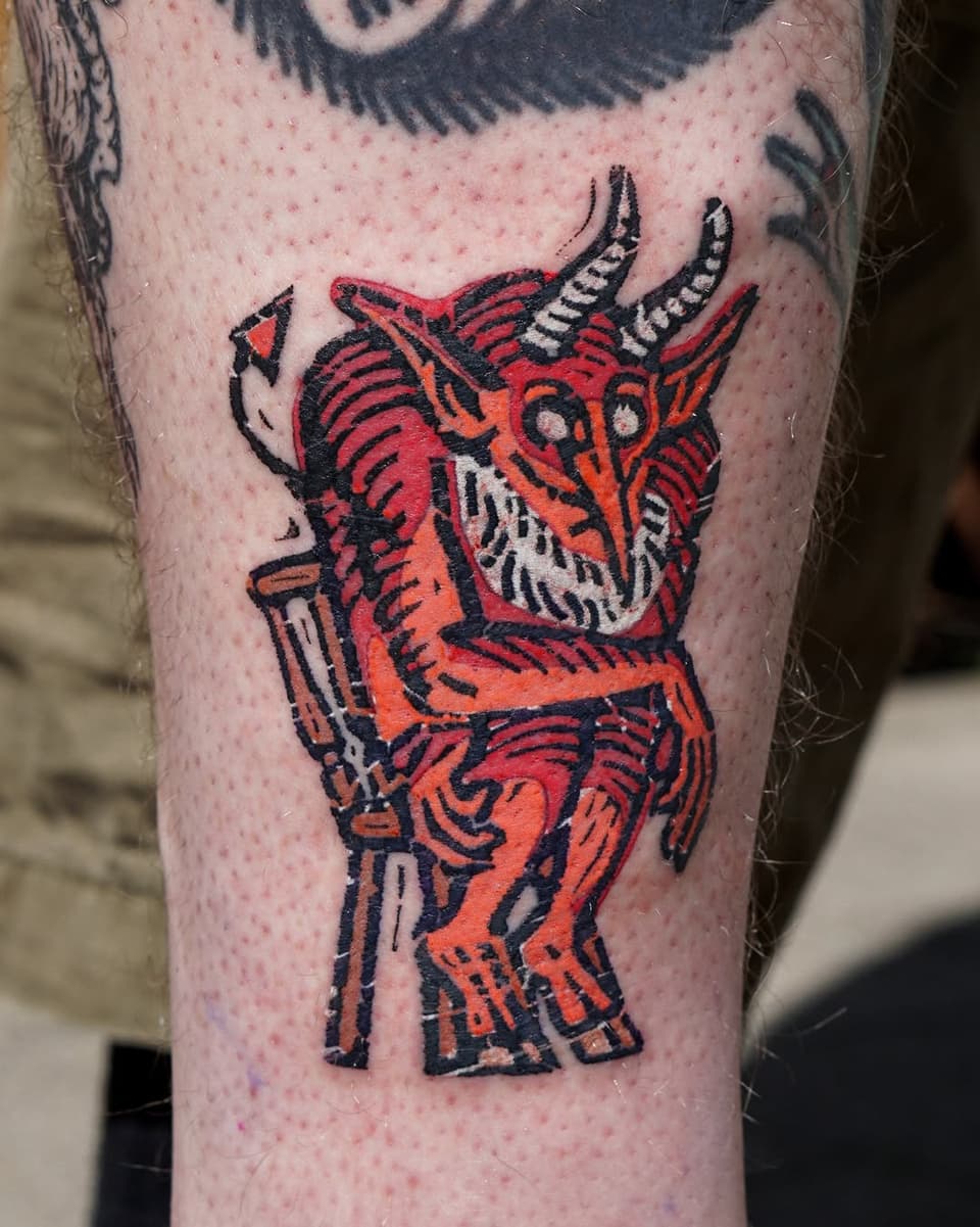 Red and orange demon creature with curved horns and a trident, featuring black outlines and white details, tattooed on the thigh.