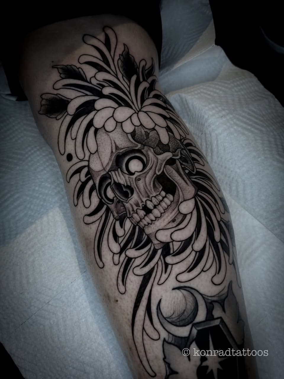 Ornate skull tattoo on the calf featuring flowing organic leaf and feather motifs in black ink with gray shading.