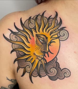 Colorful shoulder tattoo featuring a sun-faced lion with flaming mane in yellow and orange, surrounded by decorative cloud scrolls in grey and black.
