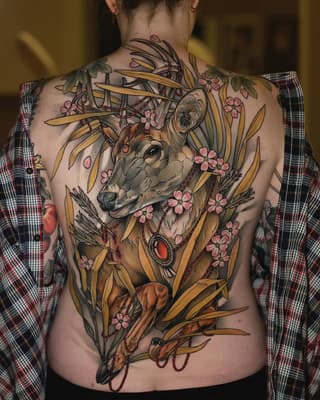 Back tattoo featuring a deer head with antlers surrounded by golden wheat stalks, pink flowers, and red gemstone accents in warm earth tones.