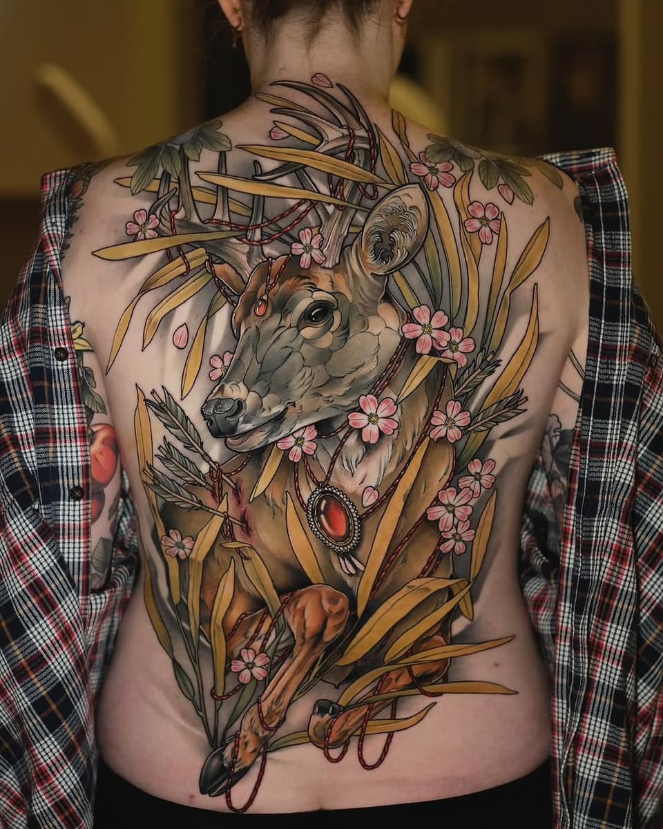 Back tattoo featuring a deer head with antlers surrounded by golden wheat stalks, pink flowers, and red gemstone accents in warm earth tones.