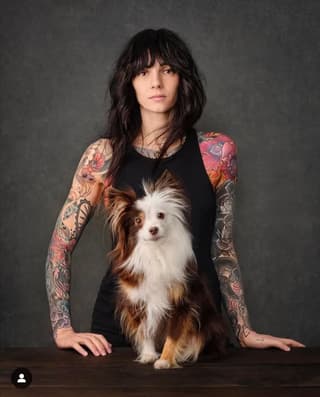 Woman with full sleeve tattoos featuring colorful floral and ornamental designs on both arms, posing with a small brown and white dog.