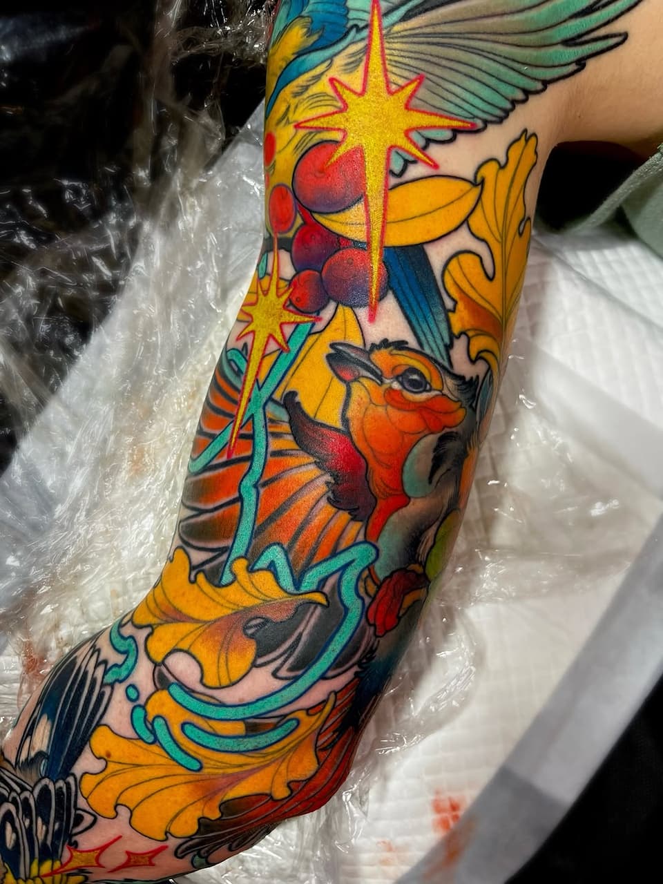 Colorful full-length sleeve tattoo featuring winged creatures, stars, ornamental scrollwork, and botanical elements in yellow, red, blue, and orange on the arm.