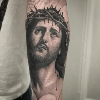 Monochromatic portrait tattoo on forearm depicting a bearded man's face wearing a crown of thorns with dripping details.