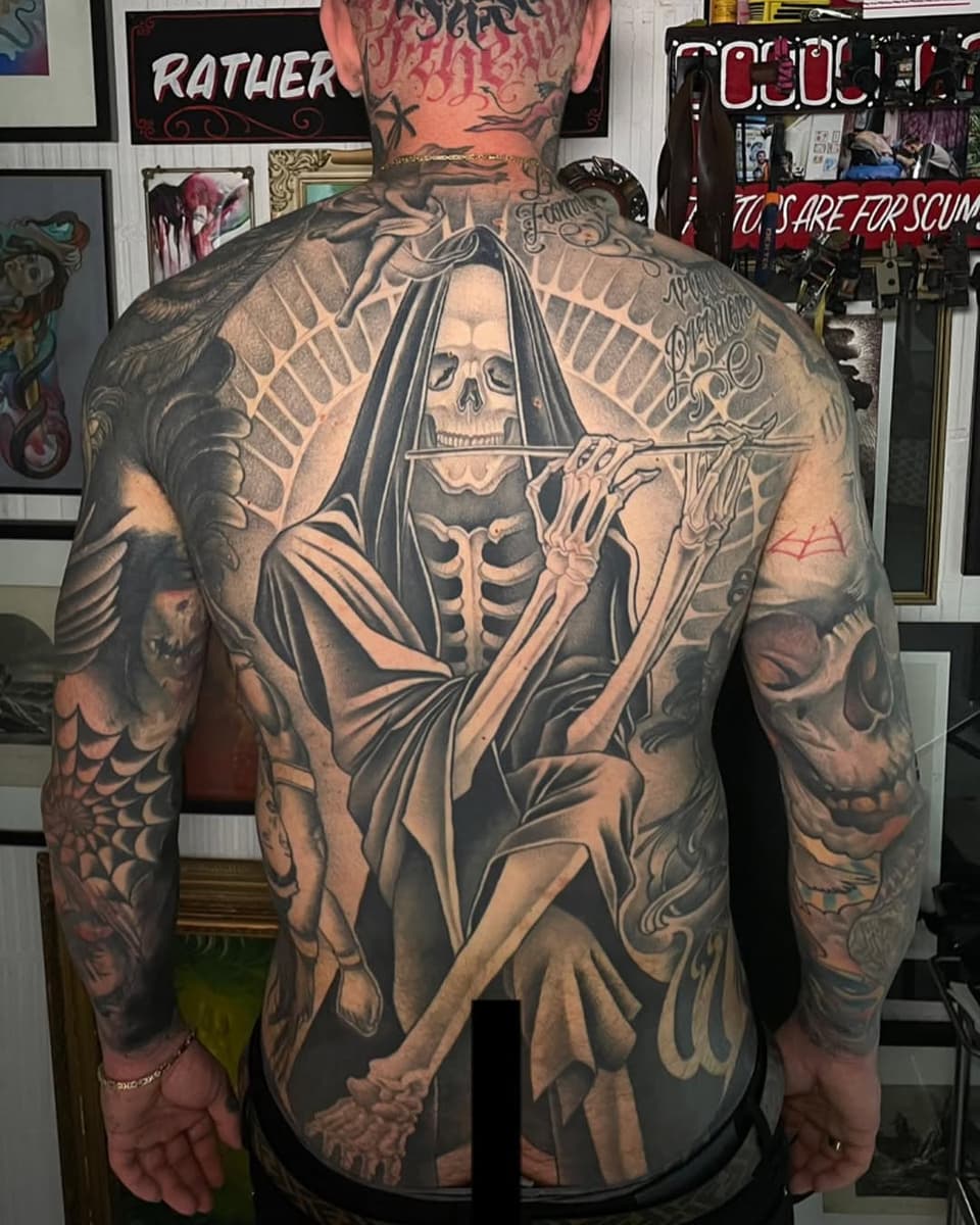 Back tattoo featuring a skeletal figure in robes holding a scythe, surrounded by skull imagery and decorative elements in black and grey ink.