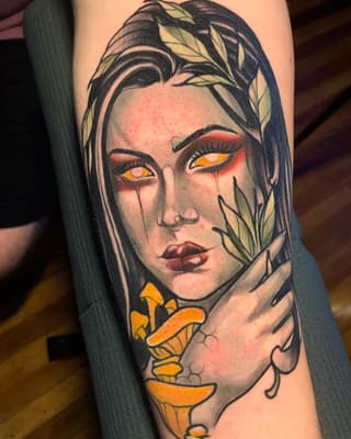 Colorful portrait tattoo on forearm featuring a woman's face with glowing orange eyes, tears, leafy hair, and golden flowers near her chin.