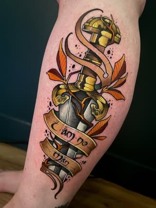 Colorful leg tattoo featuring stacked ornate armor pieces with gold and silver accents, orange leaves, brown ribbons with symbols, and decorative swirls.