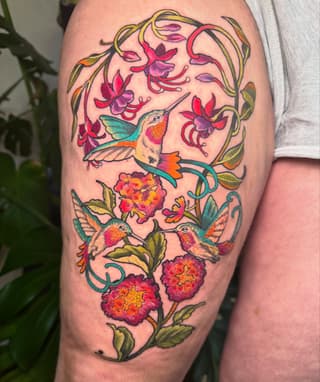 Colorful hummingbird tattoo on upper arm surrounded by pink and red flowers, green leaves, and curved vines in vibrant reds, oranges, teals, and golds.