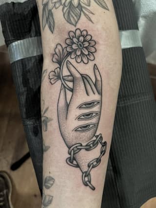 Tattoo on forearm featuring a stylized face with three eyes holding a daisy flower, wrapped in chain or rope imagery, rendered in black ink.