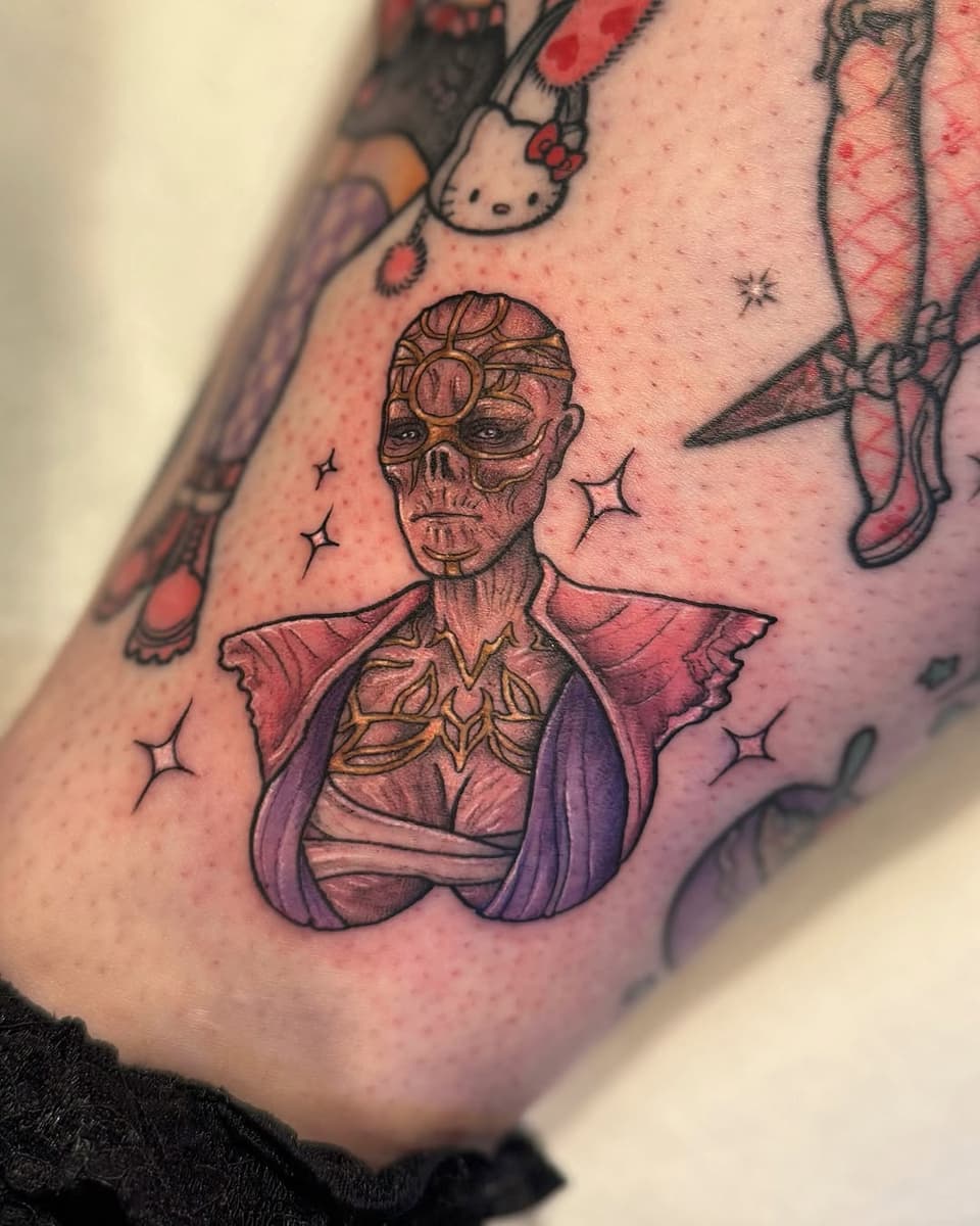 Skeletal figure with ornate facial markings and purple cape adorned with gold details, surrounded by sparkles, tattooed on the arm.