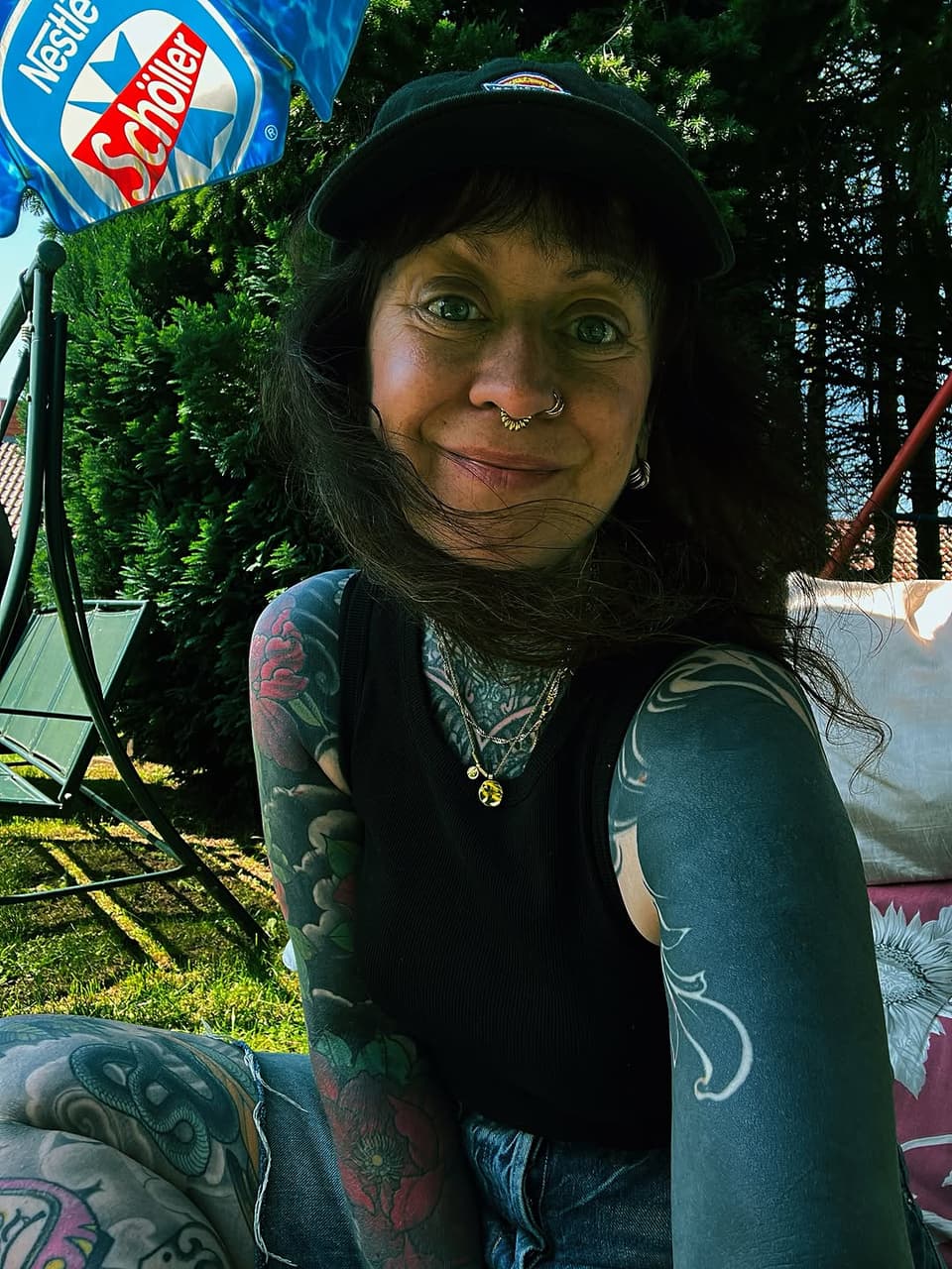 Woman with extensive arm and leg tattoos featuring colorful designs in green, red, and black displayed outdoors in natural sunlight.