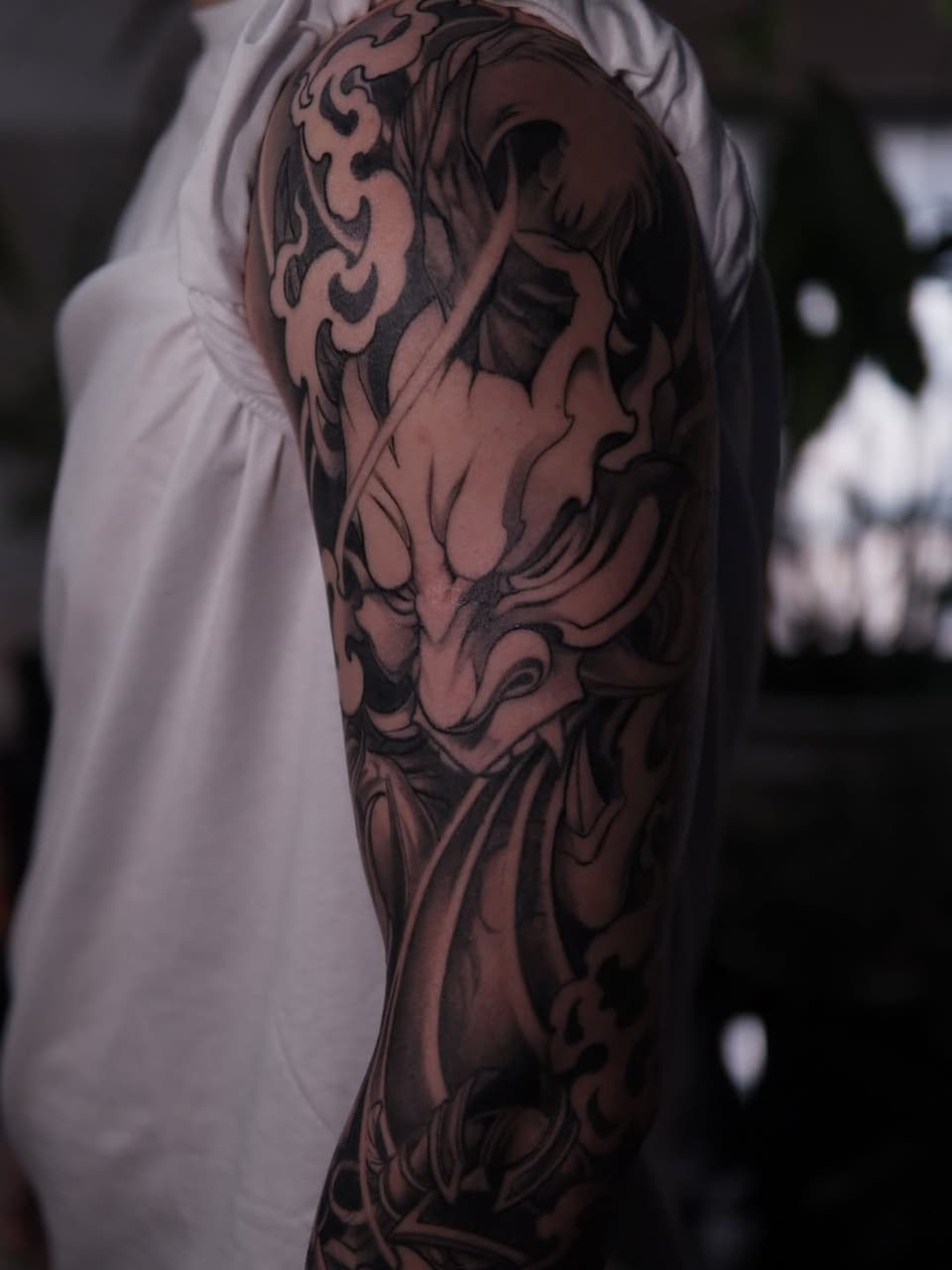 Detailed demon or oni mask tattoo on upper arm and shoulder with intricate line work and shading in dark ink on skin.