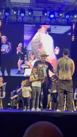Large-scale back tattoo featuring intricate golden and dark designs displayed on stage during a tattoo convention presentation with audience members present.