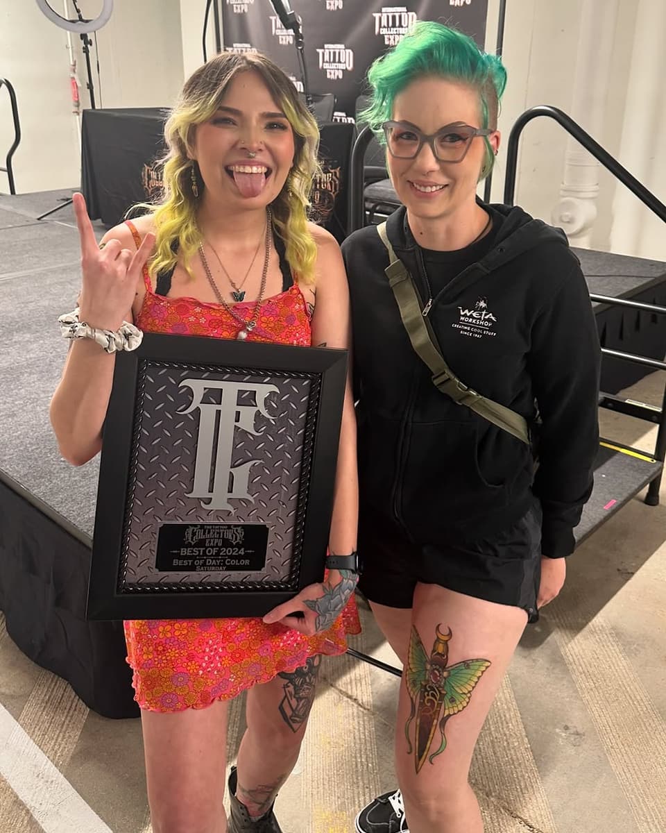 Two people posing together indoors, the person on the left holding an award plaque and wearing a red sequined dress, both displaying visible tattoos on their legs featuring insects and botanical elements in color.