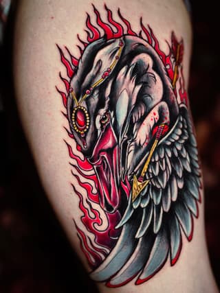 Colorful tattoo on upper arm featuring a skull with wings, surrounded by red flames and ornamental gold jewelry details.