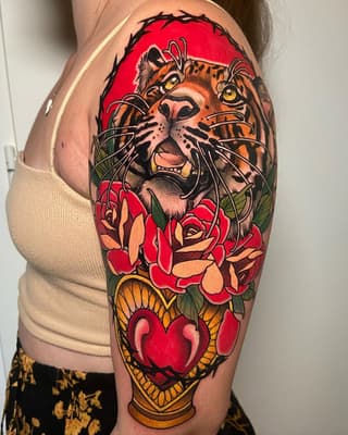 Colorful shoulder tattoo featuring a roaring tiger's head with yellow eyes, red roses, green leaves, and a red heart in a decorative frame against a bright red background.
