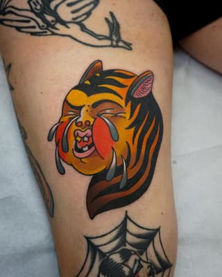 Colorful tiger head tattoo on thigh featuring orange, yellow, black and red tones with expressive facial features and striped mane.