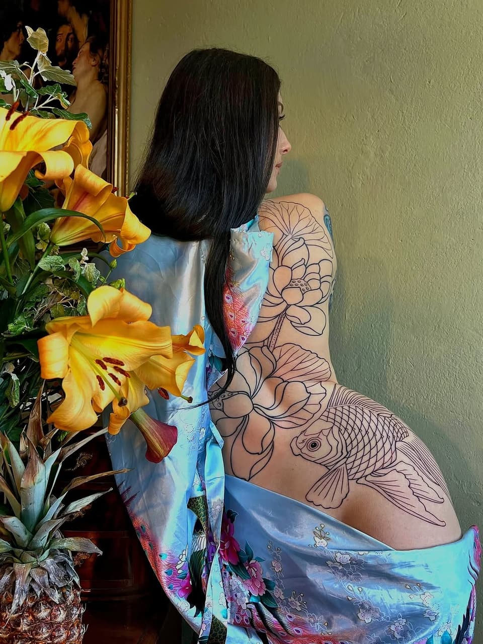 Woman displaying a large floral and koi fish tattoo in black outline covering her entire right shoulder and back in profile view.