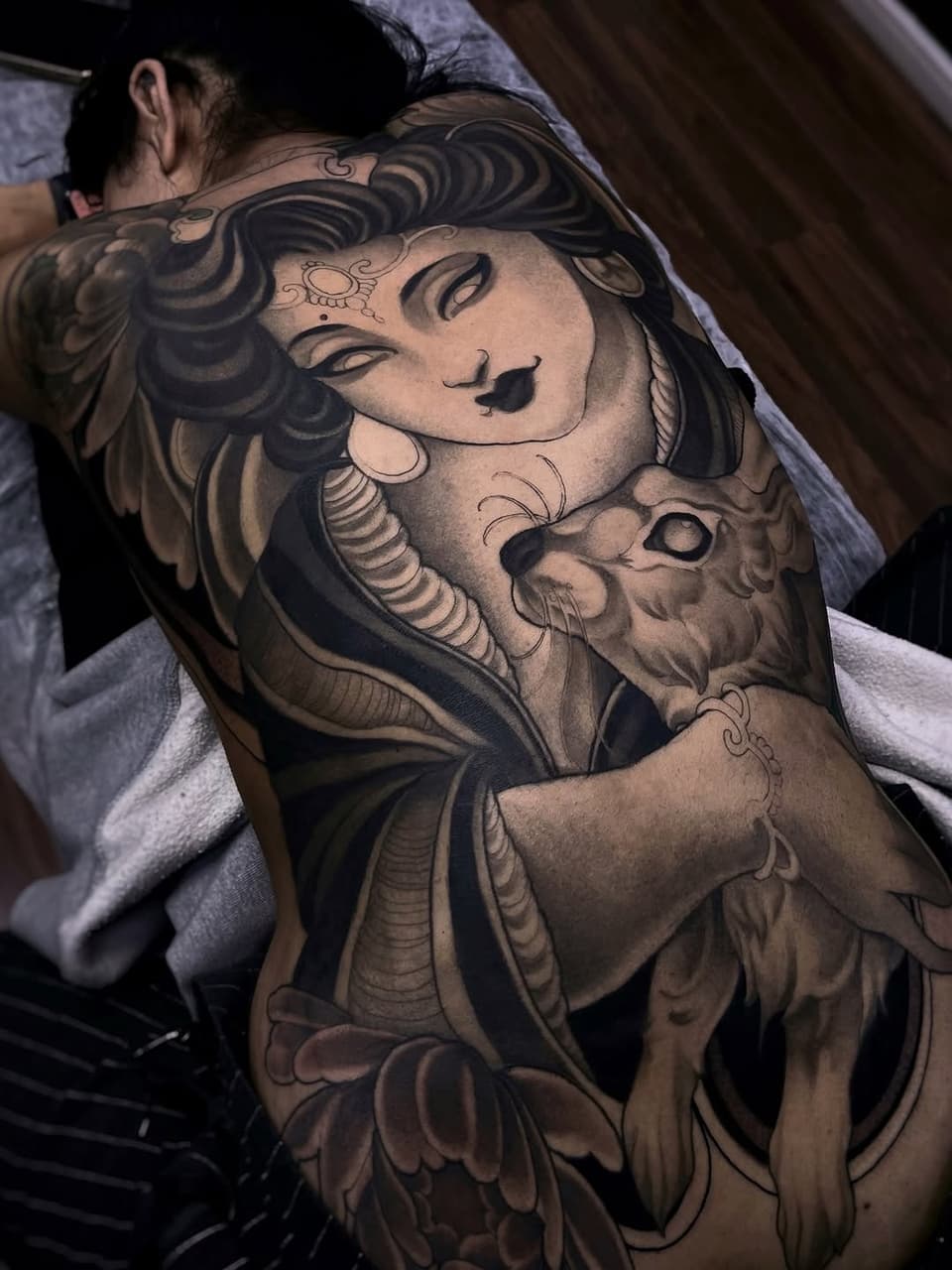 Large back tattoo featuring a woman's face with closed eyes and decorative swirls, paired with a lion head below in black and grey ink.