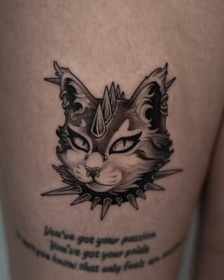 Decorative cat face with pointed ears, mystical markings, spiked collar, and text below on upper arm in black ink.