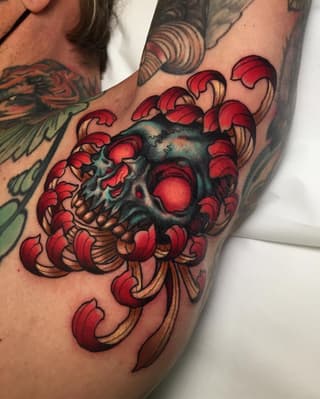 Colored skull tattoo surrounded by red ornamental scrollwork and flourishes on the upper arm, featuring teal and red shading.