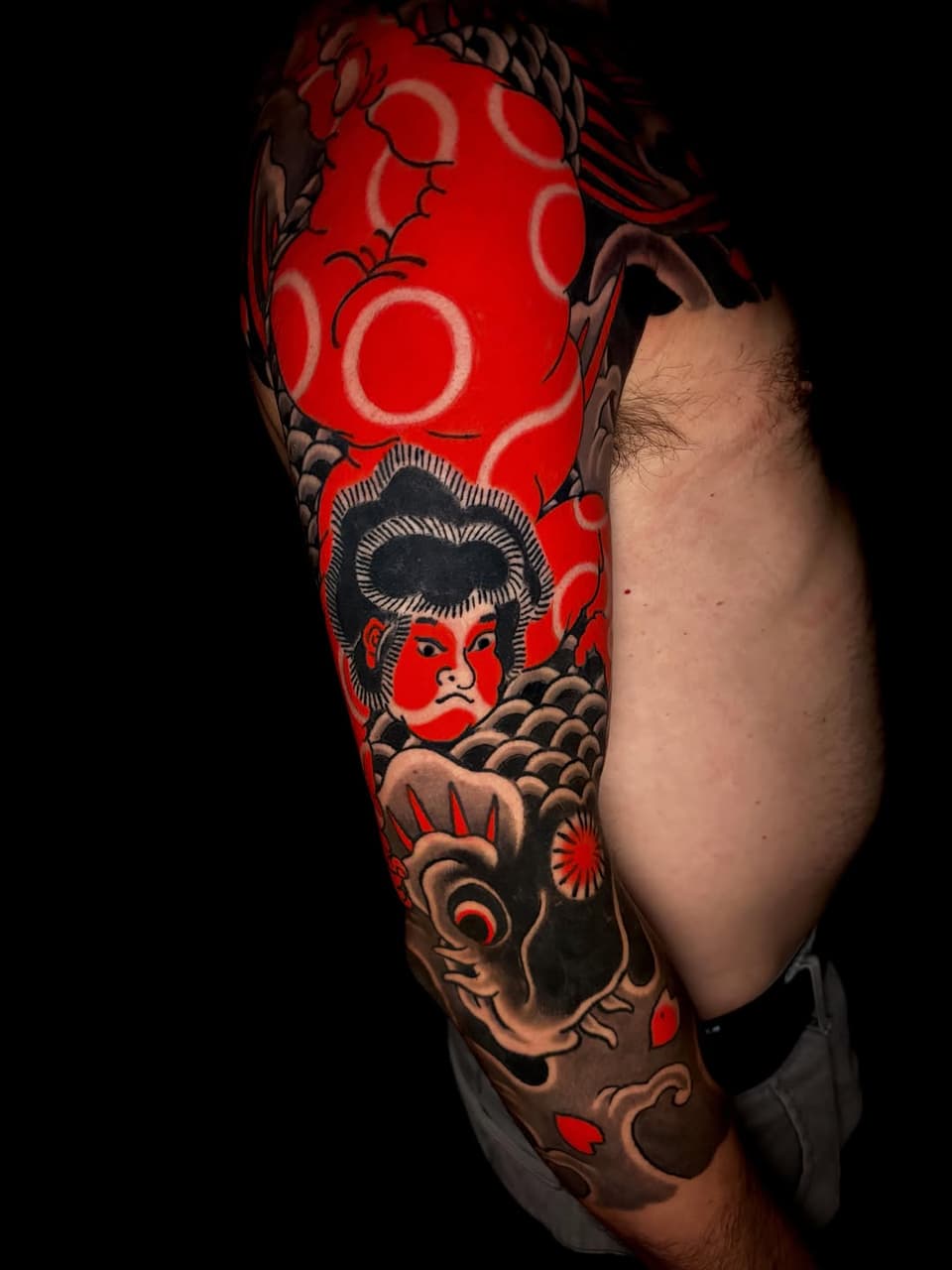 Full-sleeve arm tattoo featuring a portrait with dark hair and red face, circular patterns, a demon mask, and decorative elements in red, black, and tan tones.