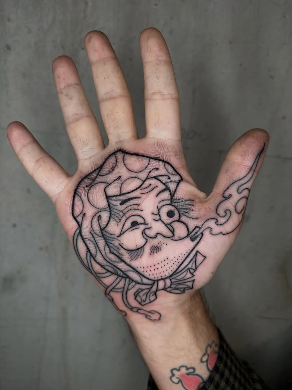 Smiling bearded face with ornate hair and curving details tattooed on open palm in black ink.