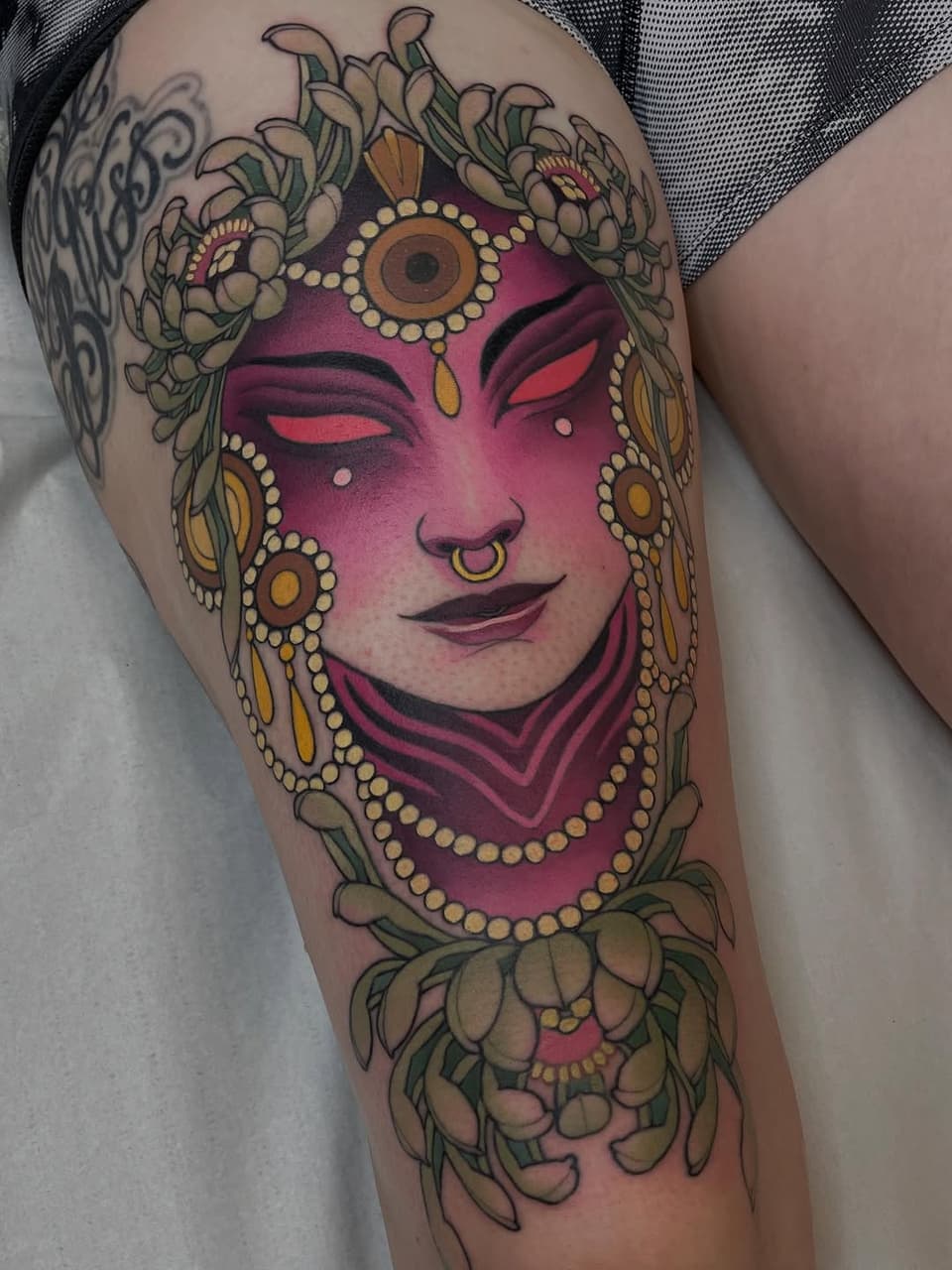Colorful tattoo on thigh featuring a magenta-faced figure with gold jewelry, ornamental flowers, and decorative elements surrounding the face.