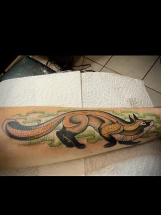 Colorful weasel or ferret tattoo on forearm featuring brown, tan, black, and green tones with grass background detail.