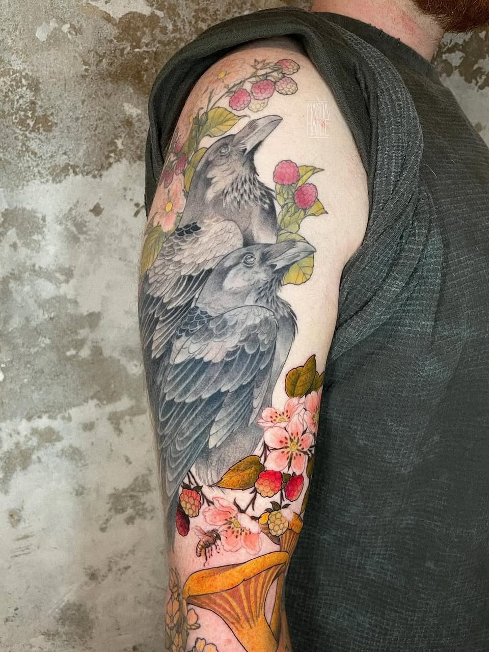 Two detailed birds with outstretched wings surrounded by pink raspberries, yellow leaves, and pink flowers on a full-sleeve arm tattoo in color and grayscale.