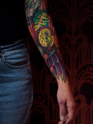 Colorful parrot tattoo on forearm featuring yellow, green, purple, and red hues with floral elements above, displayed on a person wearing denim.