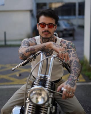 Tattooed man with colorful sleeve tattoos on both arms posing on a motorcycle, wearing sunglasses and a cream-colored shirt.