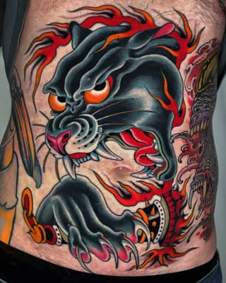 Demonic face tattoo with glowing orange eyes, red flames, sharp teeth, and decorative elements in black, red, orange, and gray on the chest.