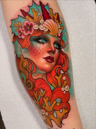 Colorful portrait tattoo of a woman's face with turquoise eyes, red hair, coral skin tones, and an elaborate headdress featuring flowers, pearls, and ornamental details on the forearm.