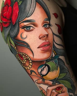 Colorful portrait tattoo on arm featuring a woman with dark hair, red flowers, a small bird, crescent moon marking, gold beaded necklace, and red petals.