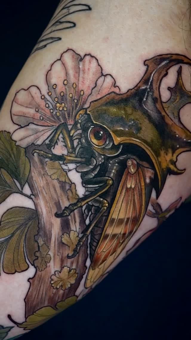 Detailed bird head tattoo on forearm with pink flowers, golden wings, and wood-grain background in brown, green, and rust tones.