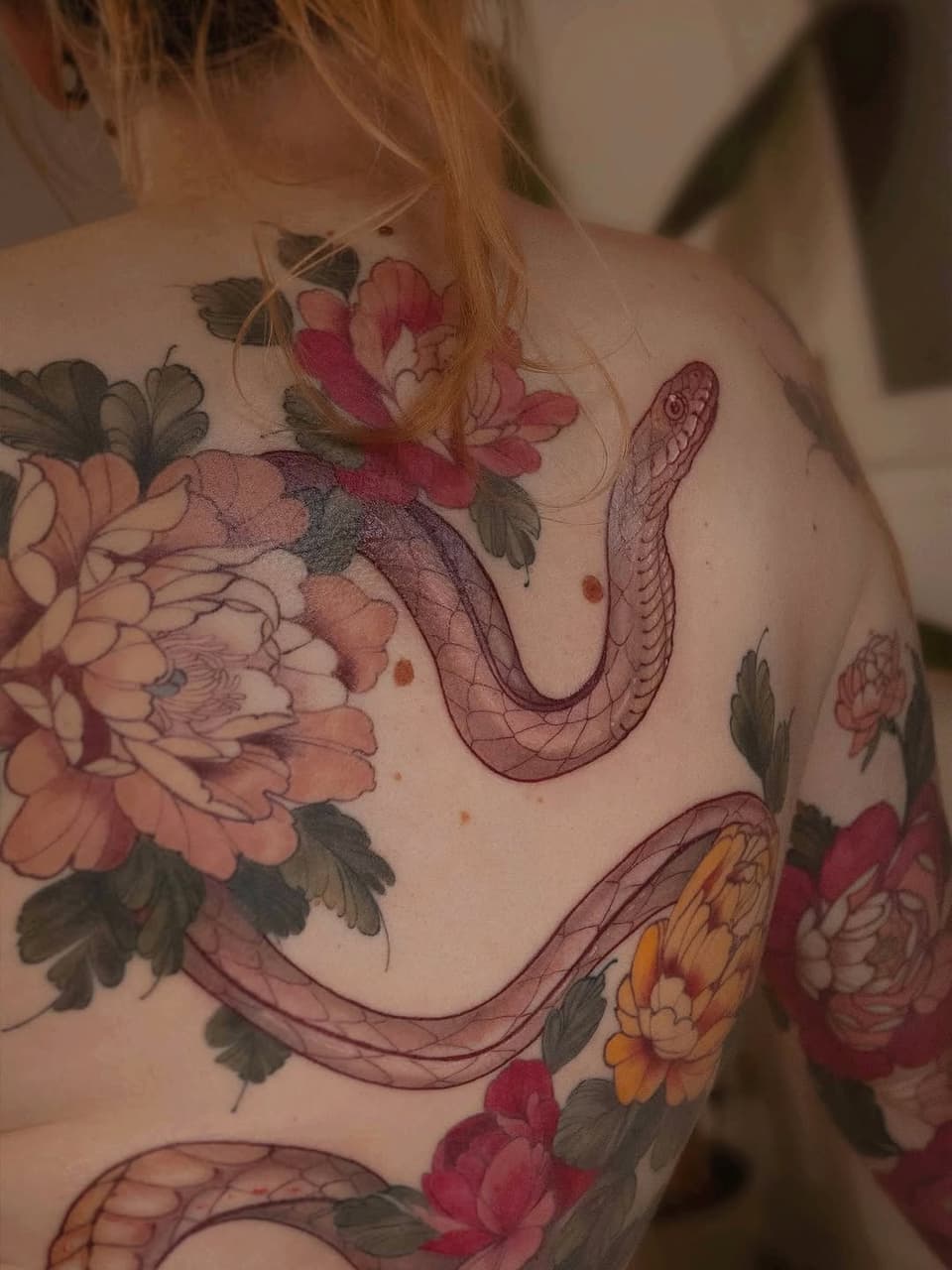 Colorful floral and serpent tattoo covering the shoulder and upper back, featuring peonies, roses, and intertwining snakes in red, pink, yellow, and green tones.